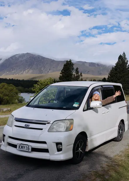 Smiling traveller waving from a white campervan with mountain views behind